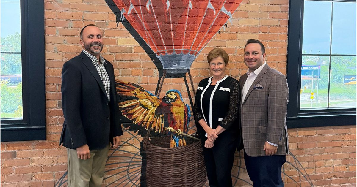 Photo of three people in suits in front of an exposed brick wall with a mural of a hot air balloon painted on it as Nancy Catarisano welcomes Cause + Effect Strategy to Insero Advisors